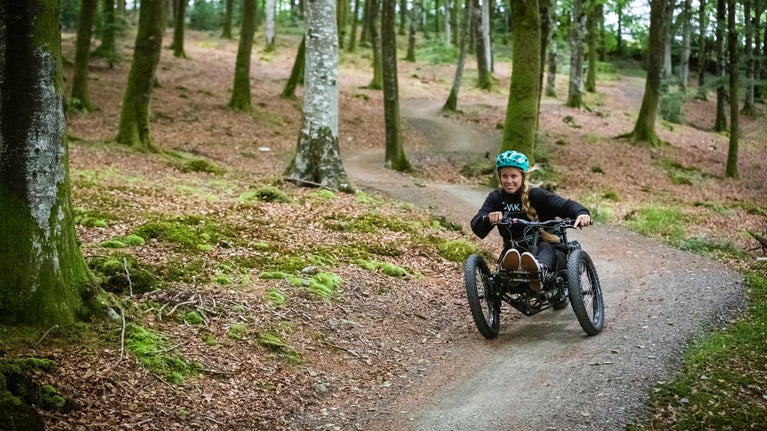 A visitor cycling using an adapted bike on a woodland trail at Lanhydrock. They cycle towards the camera on a curved trail that goes between tall trees with the woodland floor covered in dried leaves and moss.
